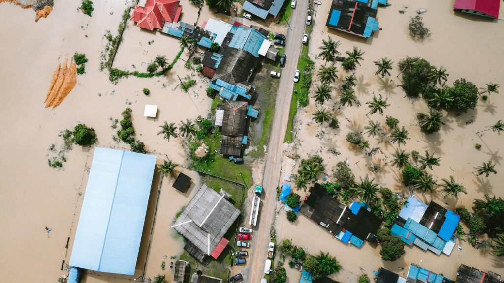 Aerial view of an area flooded with light brown water with roofs of houses, palm trees and farm buildings visible on the sides of the road dotted by cars on each side.